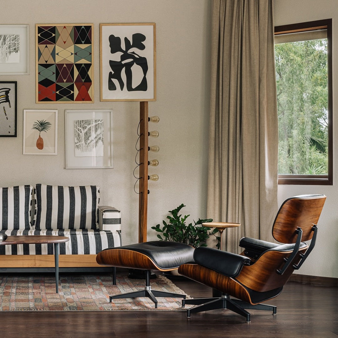 A mid-century modern lounge chair with a matching ottoman in brown wood and black leather, styled in a contemporary living room next to a striped sofa and framed wall art.