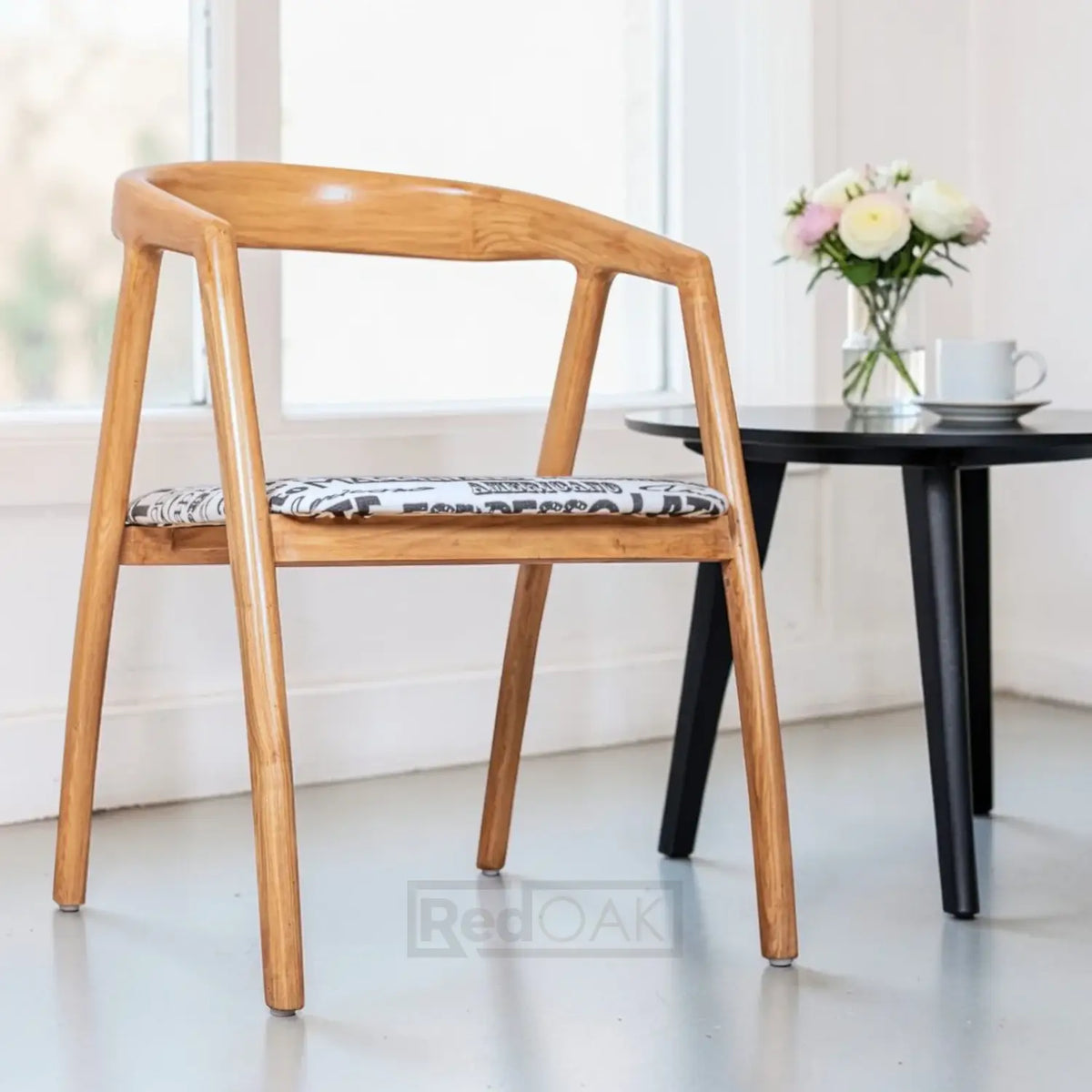 Wooden chair with a patterned cushion in a bright room with a table and flowers.