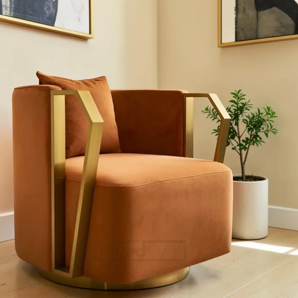 A modern accent chair in warm rust-orange velvet upholstery with geometric gold metal frame accents and a matching cushion, shown in a styled interior setting with a potted plant and framed wall art.